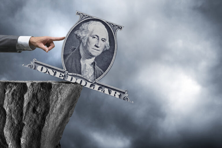 A hand in a business suit is lightly pushing a dollar bill over a cliff.